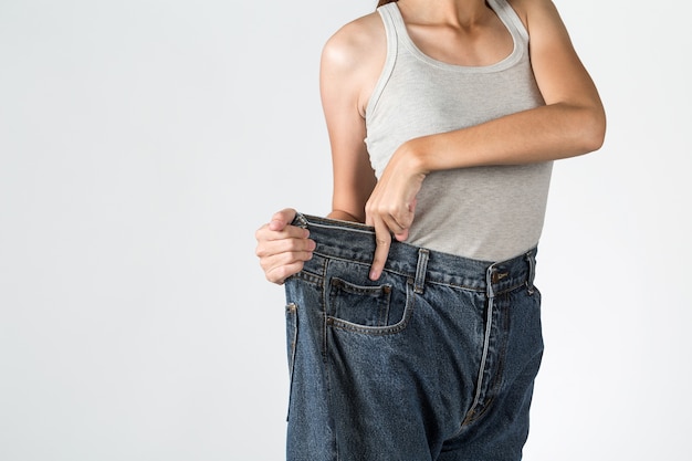 Junge Frau zeigt Gewichtsverlust mit großen Jeans und zeigt die Hand, die an den Jeans geschnitten wird