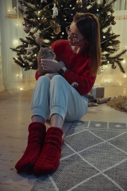 Junge Frau zusammen mit einer Katze, die nahe dem Weihnachtsbaum sitzt Weihnachten mit einer Hauskatze