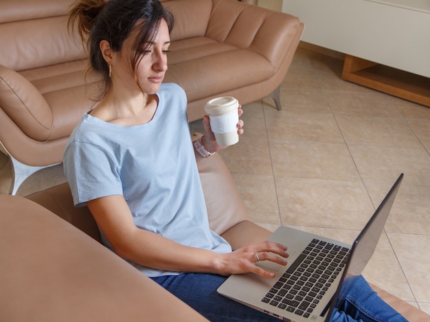 Junge Frauen, die auf dem Laptop mit Kaffee tippen, um in die Hand zu gehen, während sie zu Hause auf dem beigefarbenen Sofa sitzen