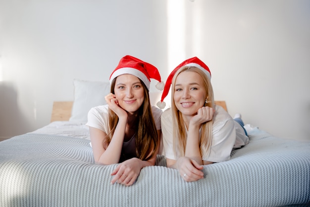 Junge Frauen, die Weihnachtsmütze tragen und lächeln. Zwei schöne Mädchen, die auf einem Bett liegen. Lächelnde und plaudernde Mädchen. Horizontale Ansicht Freundschaft und Feierkonzept.