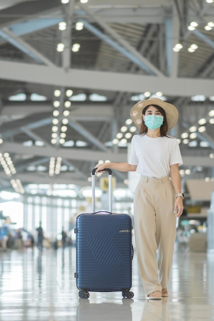 Junge Frauen mit Gesichtsmaske und Gepäck, die im Flughafenschutz spazieren gehen Coronavirus-Krankheitsinfektion Asiatische Reisende mit Hut Reisezeit nach dem Konzept der Impfstoff-Booster-Dosis