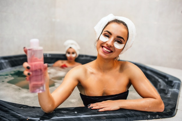 Junge Frauen sitzen im Hydromassagebad und schauen gerade. Sie hat eine Flasche Duschgel in der Hand. Ihre asiatische Freundin sitzt hinten im Whirlpool.