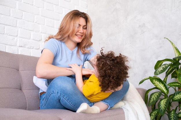 Junge Frauen spielen mit ihrer lockigen Tochter auf der Couch, sie lachen, umarmen sich und haben Spaß zusammen