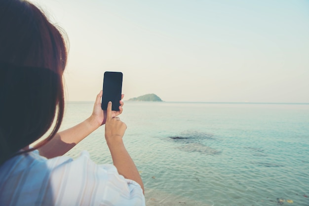 Junge Frauenhände benutzen Smartphone, das auf einem Felsen sitzt und den Strand genießt