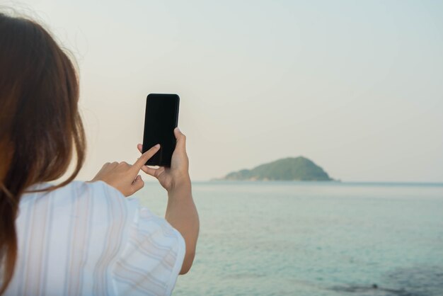 Junge Frauenhände benutzen Smartphone, das auf einem Felsen sitzt und den Strand genießt