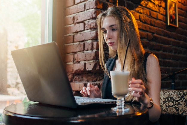 junge Freiberuflerin, die Kaffee trinkt, der auf Laptop in einem modischen Restaurant arbeitet