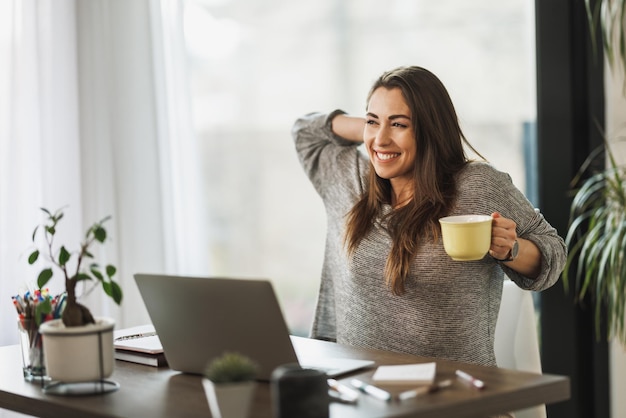 Junge Freiberuflerin genießt eine Tasse Kaffee, während sie von ihrem Heimbüro aus arbeitet.