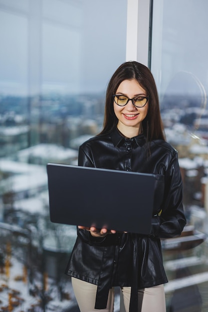 Junge Freiberuflerin mit Brille und Laptop, die remote in einem modernen Arbeitsbereich mit großen Fenstern arbeitet Remote-Arbeit