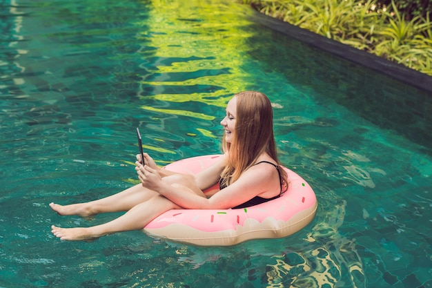 Junge Freiberuflerin sitzt am Pool mit ihrem Laptop im Hotel und surft in ihrem Smartphone. In den Ferien beschäftigt. Fernarbeitskonzept. Kopieren Sie Platz für Ihren Text.