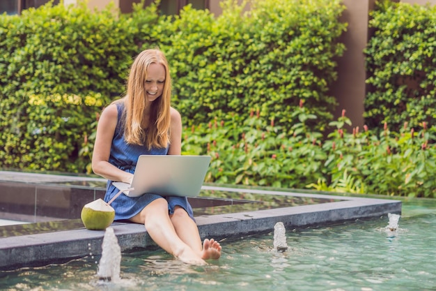 Junge Freiberuflerin sitzt am Pool mit ihrem Laptop im Hotel und surft in ihrem Smartphone. In den Ferien beschäftigt. Fernarbeitskonzept. Kopieren Sie Platz für Ihren Text.