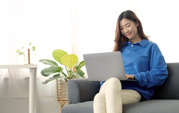 Junge, freiberuflich tätige Asiatin, die am Laptop arbeitet und soziale Medien überprüft, während sie auf dem Sofa liegt, wenn sie sich im Wohnzimmer zu Hause entspannt
