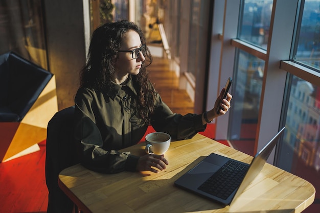 Junge freiberufliche Frau Junge Frau, die mit Laptop im Coworking Space arbeitet und am Telefon spricht, sitzt im offenen Raum mit einer Tasse Kaffee und lächelt, während sie eine Brille hält