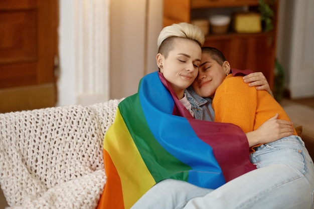 Junge freiheitliche lesbische Frau kuschelt in LGBT-Regenbogenfahne, während sie zu Hause auf dem Sofa im Wohnzimmer sitzt