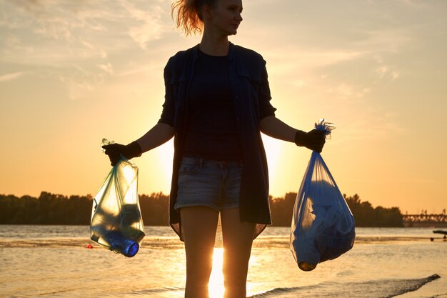 Junge Freiwillige in schwarzen Handschuhen geht mit Müllsack an einem schmutzigen Strand des Flusses entlang