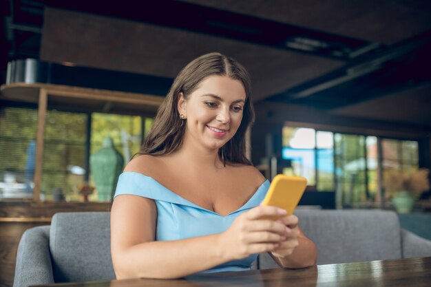 Junge freudige lächelnde Frau, die interessiert an Smartphone sitzt, das im Sommercafé sitzt