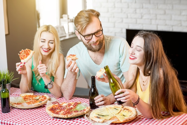 Junge Freunde, die lässig in bunten T-Shirts gekleidet sind, essen zu Hause mit Pizza und Bier zu Mittag