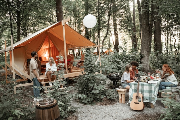 Junge Freunde haben Spaß beim Glamping nach Sonnenuntergang. Fröhliches tausendjähriges Camping der Jugendbande bei Open-Air-Picknick unter Glühbirnenlichtern. Freundschaftskonzept mit Jungs und Mädchen, die Wein auf der Grillparty trinken