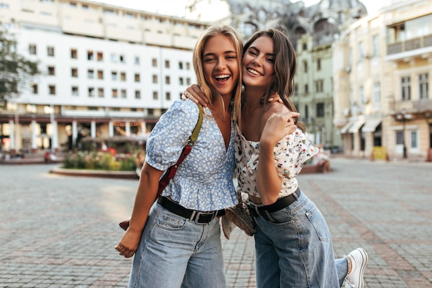 Junge Freundinnen in stylischen Jeans und floralen trendigen Blusen umarmen, lächeln und posieren gut gelaunt auf dem Stadtplatz