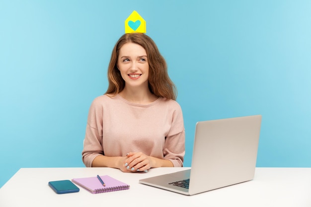 Junge, freundliche, positive Frau, die mit Laptop am Arbeitsplatz sitzt und ein kleines Papierhaus auf dem Kopf hält, träumt vom Kauf einer eigenen Wohnung, Studioaufnahme isoliert auf blauem Hintergrund