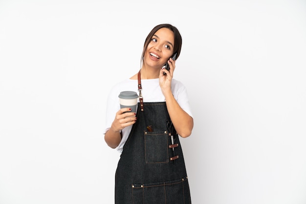 Junge Friseurfrau lokalisiert auf weißer Wand, die Kaffee hält, um und ein Handy wegzunehmen