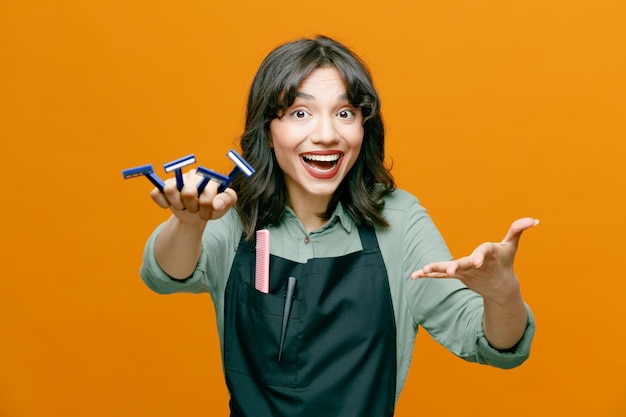 Junge Friseurfrau mit Schürze und Rasiermessern, die glücklich und positiv lächelnd in die Kamera schaut und sich fröhlich mit dem Arm präsentiert, der über orangefarbenem Hintergrund steht