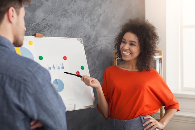 Junge fröhliche afroamerikanische Geschäftsfrau, die dem Kollegen im Sitzungssaal eine Präsentation gibt. Zwei multiethnische Partner diskutieren Verkaufschart im Büro, Kopierraum