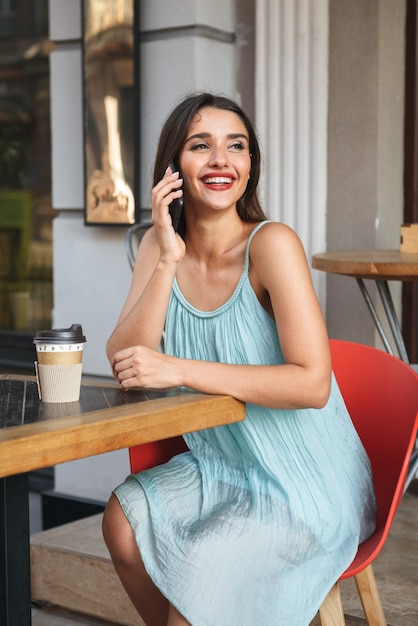 junge fröhliche Frau, die im Café draußen beim Sprechen durch das Handy sitzt, das Kaffee trinkt