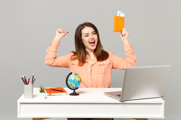 Junge fröhliche Frau, die Siegergeste macht, Erdkugel hält, Pass-Bordkarten-Ticketarbeit am Schreibtisch mit PC-Laptop isoliert auf grauem Hintergrund. Leistung Business-Karriere-Lifestyle-Konzept.