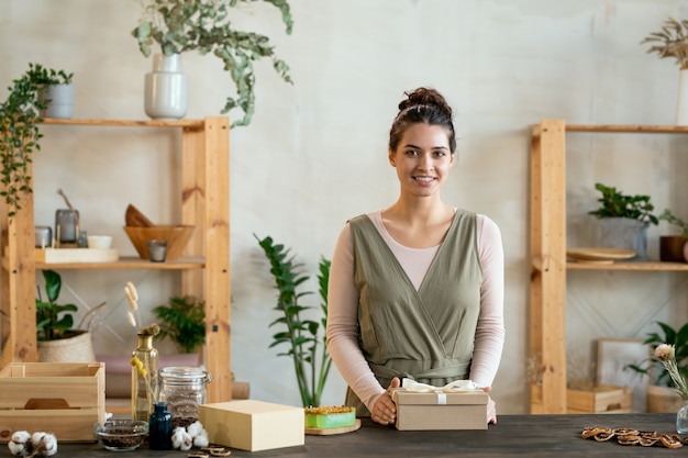 Junge fröhliche Frau in intelligenter Freizeitkleidung, die eine Geschenkbox aus Karton mit handgemachtem Geschenk hält, während sie am Tisch vor der Kamera steht