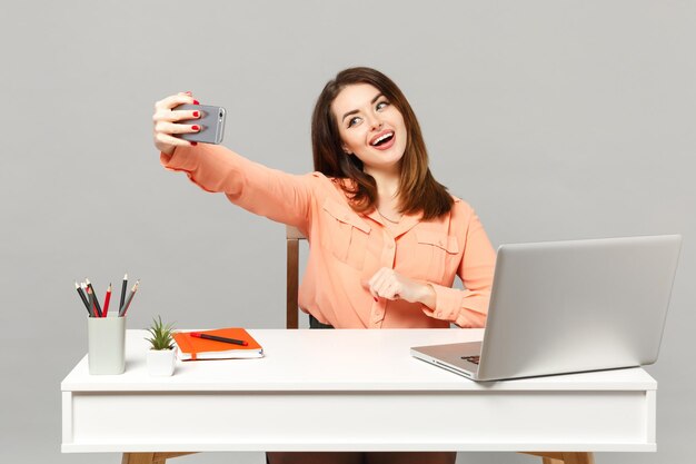 Junge fröhliche Frau in pastellfarbener Kleidung macht Selfie-Aufnahme auf dem Handy, arbeitet am Schreibtisch mit PC-Laptop isoliert auf grauem Hintergrund. Leistung Business-Karriere-Lifestyle-Konzept. Mock-up-Kopienbereich.