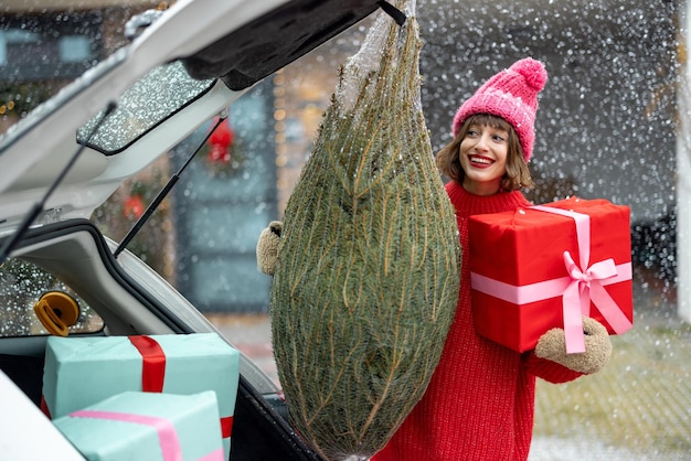 Junge fröhliche Frau in rotem Pullover und Hut mit eingewickeltem Weihnachtsbaum und Geschenkkiste in der Nähe des Autos an der Veranda ihres Hauses, dekoriert für die Winterferien Konzept der Vorbereitung auf ein neues Jahr