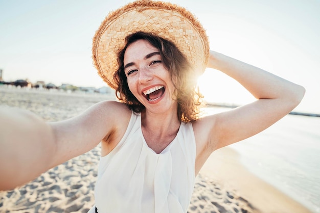 Junge fröhliche Frau in weißem Hemd mit Hut, die am Strand in die Kamera lächelt