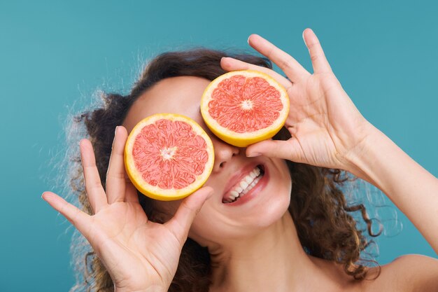 Junge fröhliche Frau mit gesunden Zähnen und Haut, die Scheiben frischer saftiger Grapefruit mit den Augen auf Blau in Isolation hält