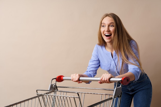 Junge fröhliche Frau mit leerem Einkaufswagen auf beigem Hintergrund