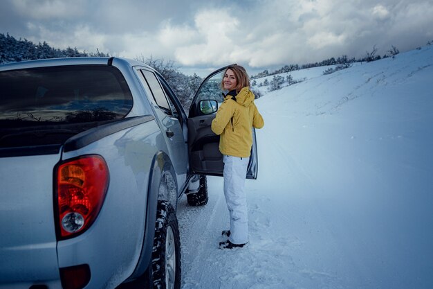 Junge fröhliche Frau posiert gegen den Auto- und Schneewald. Attraktive Frau gekleidete gelbe Jacke sitzt am Beifahrersitz im silbernen Auto, Wintersaison