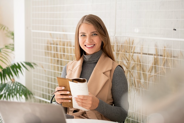 Junge fröhliche Geschäftsfrau mit dem zahnigen Lächeln, das Heft hält, während Vorbereitung für Seminar im Café