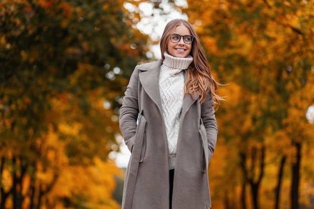 Junge fröhliche Hipsterfrau im eleganten langen Mantel mit Brille in einem gestrickten weißen Pullover steht im Park an einem warmen Herbsttag. Glückliches Mädchen genießt das Entspannen im Wald. Saisonale Damenoberbekleidung.