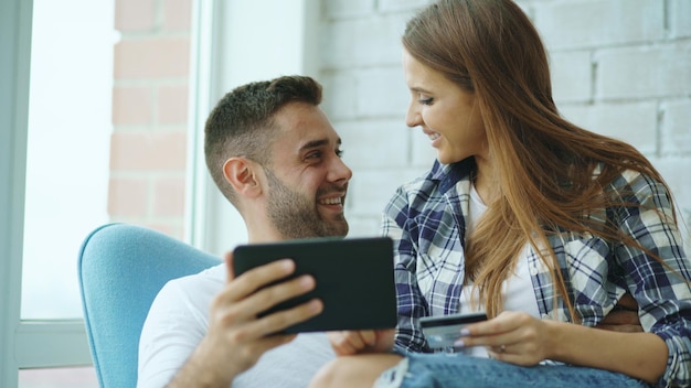 Junge fröhliche Paare, die digitale Tablet-Computer für Online-Shopping verwenden, sitzen auf dem Balkon