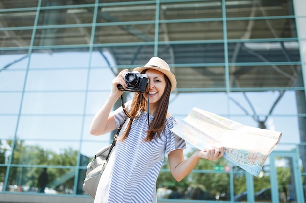 Junge fröhliche Reisende Touristenfrau fotografieren auf Retro-Vintage-Fotokamera mit Papierkarte am internationalen Flughafen