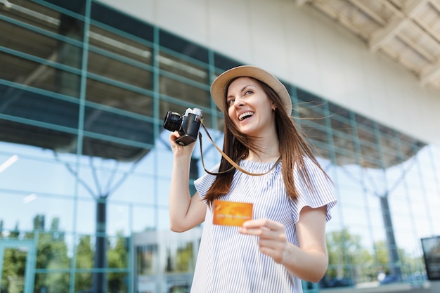 Junge fröhliche reisende touristin mit hut mit retro-vintage-fotokamera, kreditkarte am internationalen flughafen?