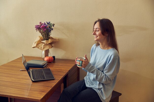 Junge fröhliche, selbstbewusste und glückliche Freiberuflerin arbeitet zu Hause in ihrer Küche am Laptop