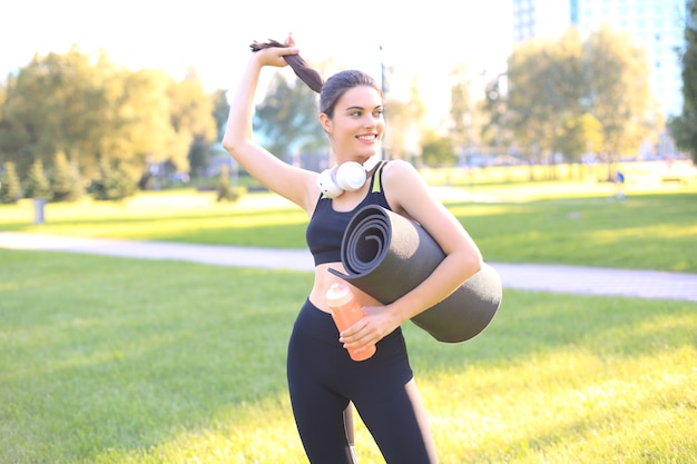 Junge fröhliche Sportfrau, die im Stadtpark mit Fitness-Teppich spazieren geht.