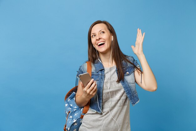 Junge fröhliche Studentin in Denim-Kleidung mit Rucksack, Kopfhörer, die Hände ausbreiten und Musik hören, halten Handy isoliert auf blauem Hintergrund. Ausbildung an der Hochschule. Kopieren Sie Platz für Werbung.