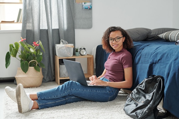 Junge fröhliche Studentin in Freizeitkleidung, die mit Laptop auf den Beinen auf dem Boden am Bett sitzt, während sie dich in der häuslichen Umgebung anschaut