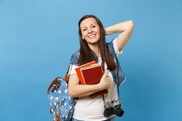 Junge fröhliche Studentin mit Rucksack und Retro-Vintage-Fotokamera am Hals, die die Hand auf dem Kopf hält, halten Schulbücher isoliert auf blauem Hintergrund. Bildung im Hochschulkonzept der High School.