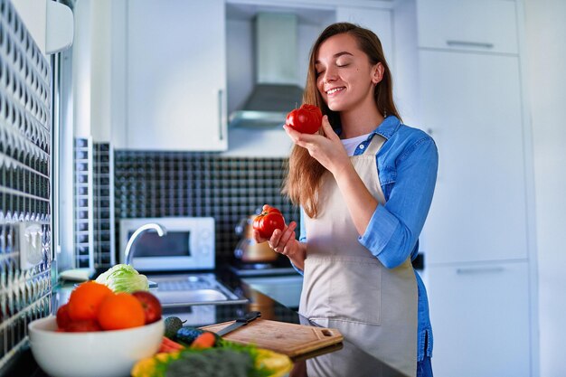 Junge, fröhliche, süße, lächelnde Hausfrau mit Schürze, die frischen vegetarischen Salat für gesundes Essen in der Küche zu Hause zubereitet