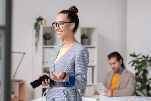 Junge fröhliche weibliche Büroangestellte mit Smartphone und Kopfhörern, die Musik genießen, während ihr Kollege Tablet hinter sich verwendet