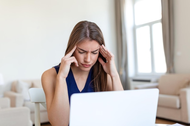 Junge frustrierte frau, die am schreibtisch vor dem laptop arbeitet und unter chronischen täglichen kopfschmerzen leidet, die online behandelt werden, um zu einer medizinischen beratung für elektromagnetische strahlung krankengeld zu ernennen