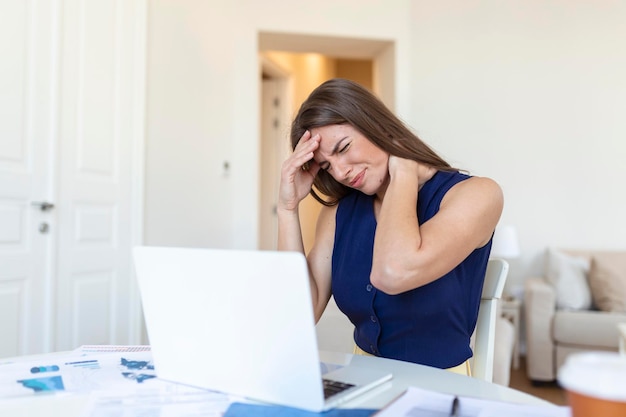 Junge frustrierte Frau, die am Schreibtisch vor Laptop arbeitet, der unter chronischen täglichen Kopfschmerzen leidet