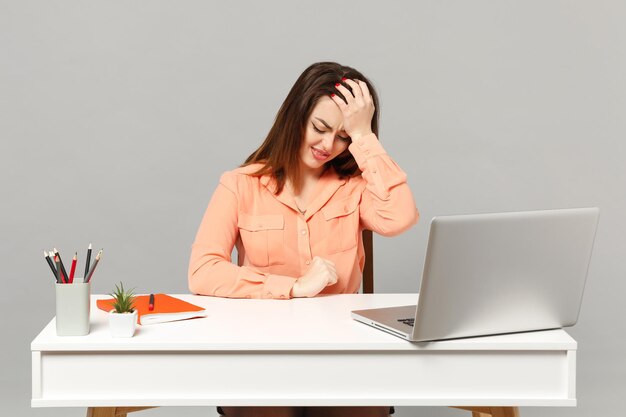 Junge frustrierte Frau in pastellfarbener Freizeitkleidung, die die Hand auf den Kopf legt, sitzt und arbeitet am Schreibtisch mit PC-Laptop isoliert auf grauem Hintergrund. Leistung Business-Karriere-Lifestyle-Konzept. Mock-up-Kopienbereich.